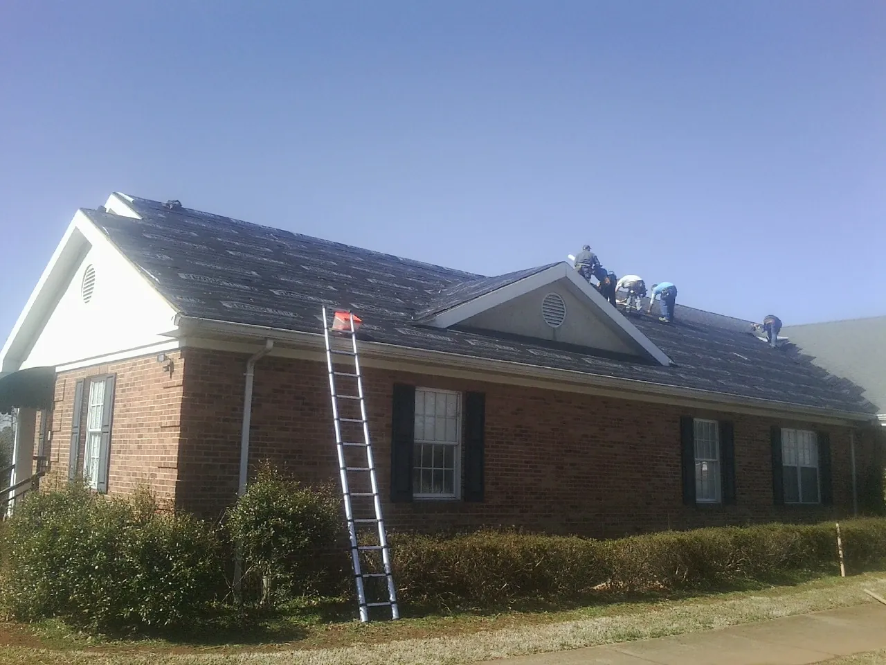 Crew tearing shingles off a brick ranch roof with a ladder set — Metal Roof Repair work by our crew in Riverdale